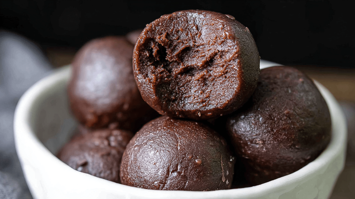 A close-up photo of several chocolate protein brownie balls stacked in a white bowl. The top ball has a bite taken out, showing the dense and fudgy interior texture.